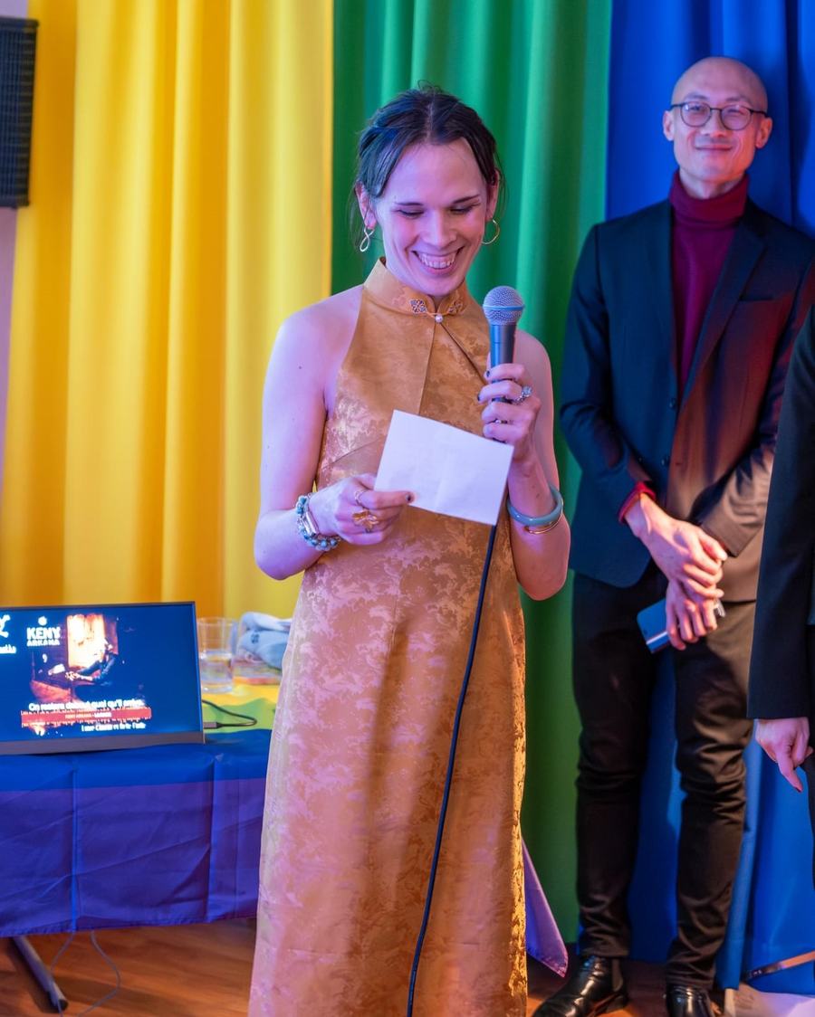 Manon Pézot in a gold dress smiling as she reads her speech into a microphone, with Olivier Li in the background