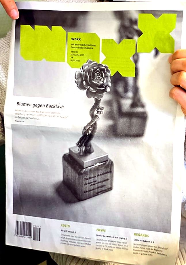 A copy of the weekly woxx held up, with a Role Models flower trophy on its cover under the headline Blumen gegen Backlash