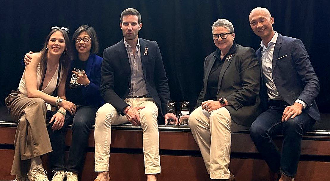 Loic Choquet and Gaby Schneider sitting on a stage edge with their crystal trophies, with members of the Blom team
